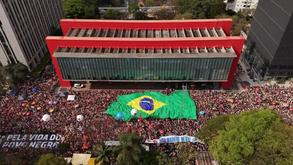 Protesto na rua com grande aglomeração de pessoas segurando bandeira do Brasil e cartazes, em área urbana com edifícios ao redor.