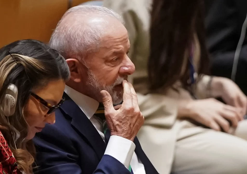Imagem de uma reunião com pessoas focadas e pensativas, incluindo um homem idoso de cabelo branco e barba, usando terno escuro, em um ambiente formal.