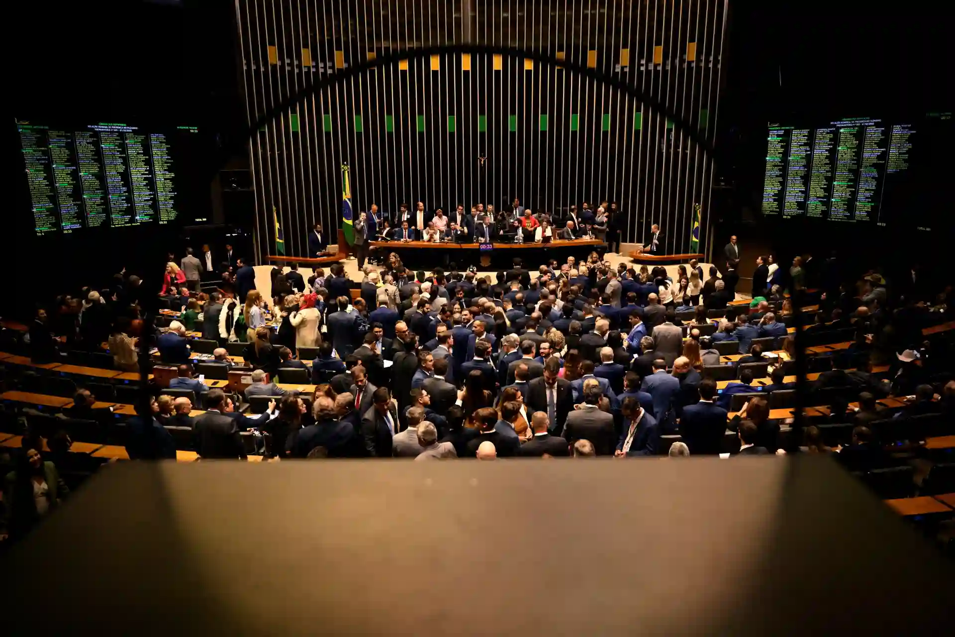Imagem de uma sessão na Câmara dos Deputados com muitas pessoas presentes, computadores e telões exibindo informações, enfatizando o ambiente político brasileiro.