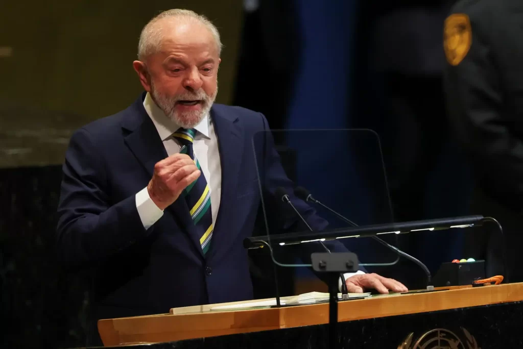 Alto funcionário falando em um púlpito, usando terno e gravata listrada, na ONU, com iluminação adequada e fundo escuro.