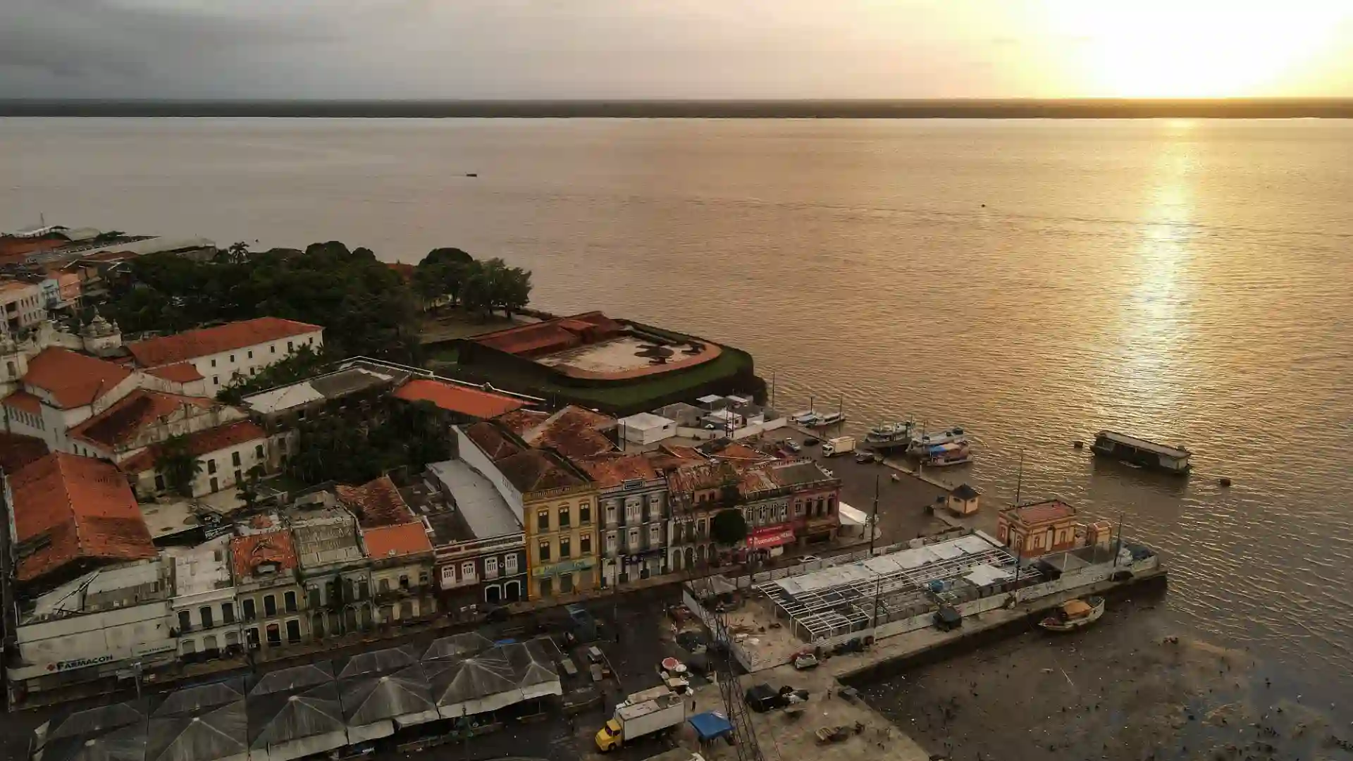 Imagem aérea de um vilarejo de hive na costa, com prédios históricos, barcos atracados e o pôr do sol ao fundo.