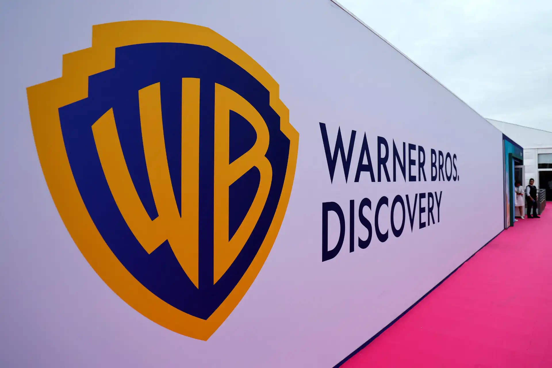 Logotipo da Warner Bros. Discovery em uma parede branca com detalhes em azul e amarelo, no evento Warner Bros. Discovery, com pessoas ao fundo.