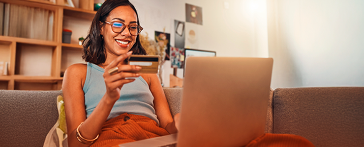 Mulher sorridente usando cartão de crédito, sentado no sofá com laptop aberto em casa, representando compras online e segurança financeira. Consumidor
