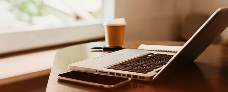 Imagem de um ambiente de trabalho com um laptop, celular, caderno e uma xícara de café sobre uma mesa de madeira, ideal para produtividade e home office.