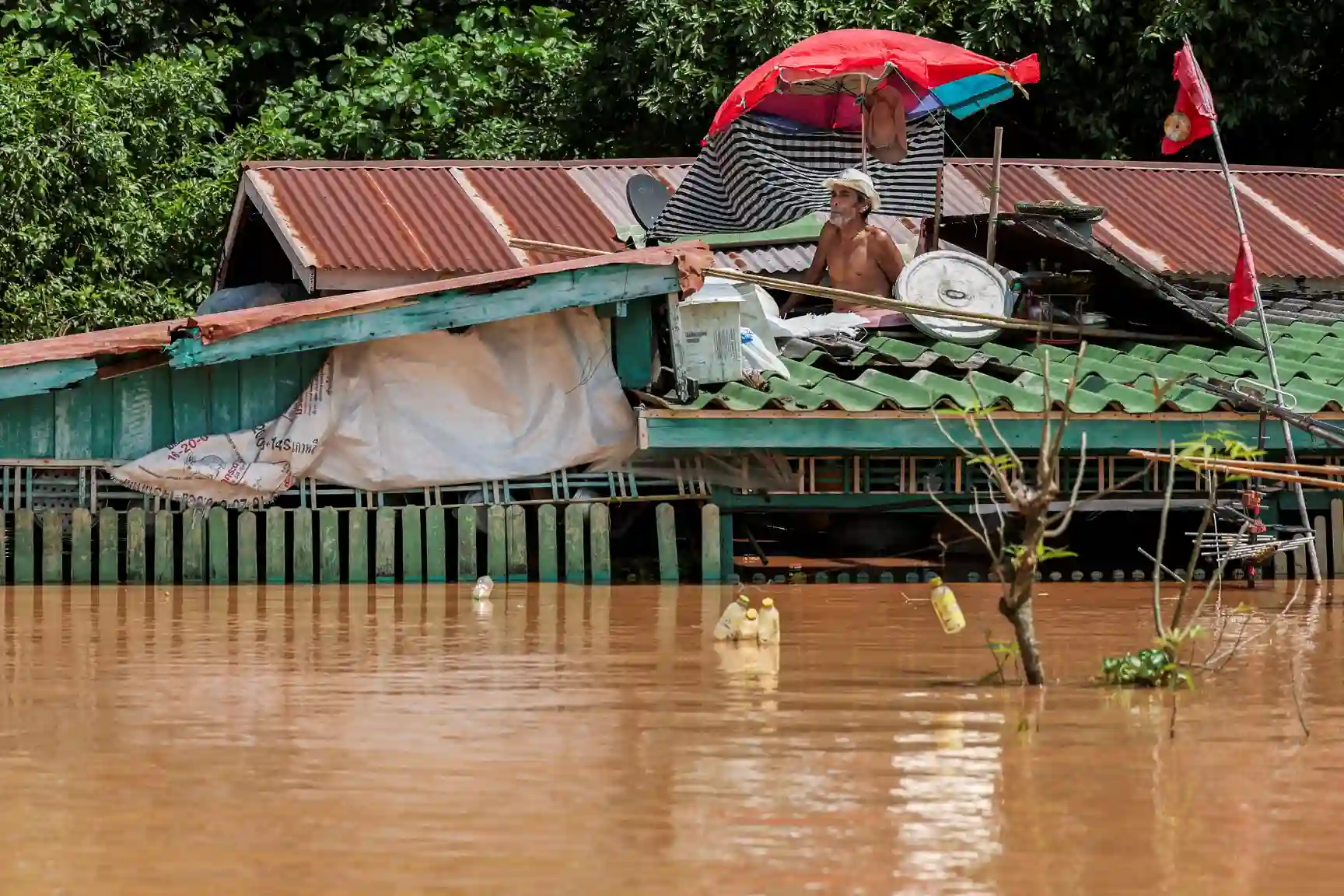 Casa de palafita alagada por enchente, com homem observando o nível da água, cercada de árvores verdes e objetos suspensos, indicando situação de emergência.