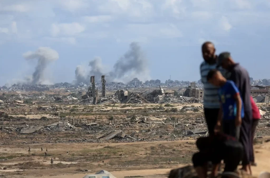 Imagem de um cenário de destruição com prédios destruídos e fumaça no horizonte, enquanto três pessoas observam, destacando o impacto da guerra.