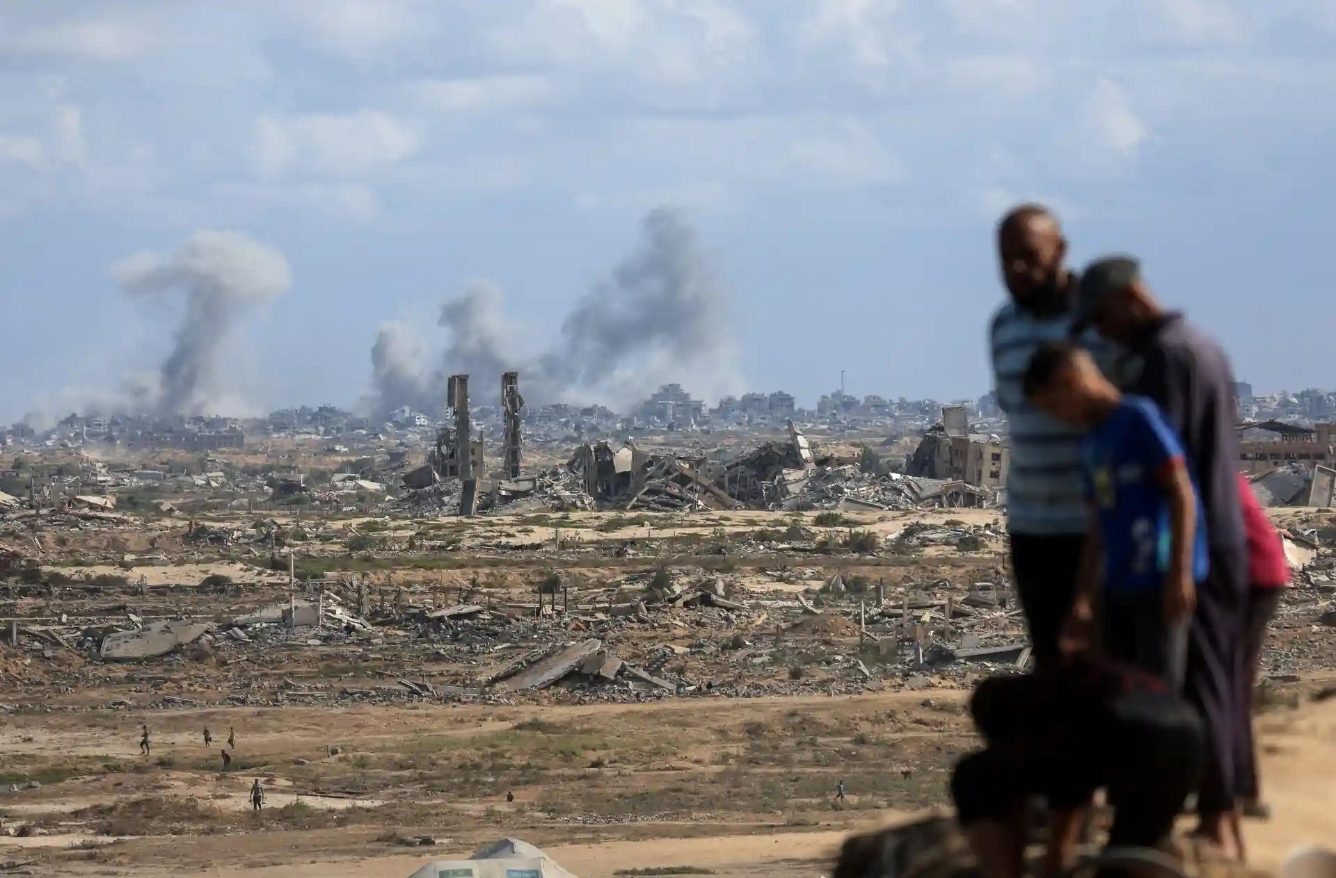 Imagem de um cenário de destruição com prédios destruídos e fumaça no horizonte, enquanto três pessoas observam, destacando o impacto da guerra.