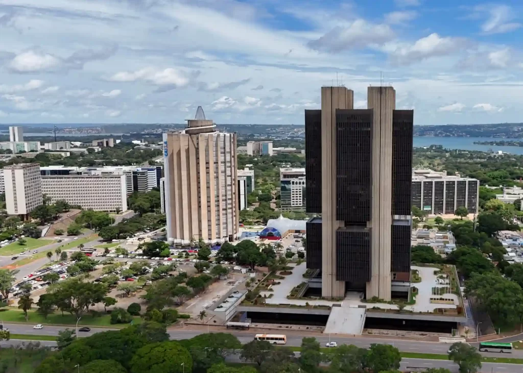 Vista panorâmica de um centro urbano com prédios elevados e áreas verdes ao redor, sob um céu parcialmente nublado, capturada de um drone ou altura elevada.