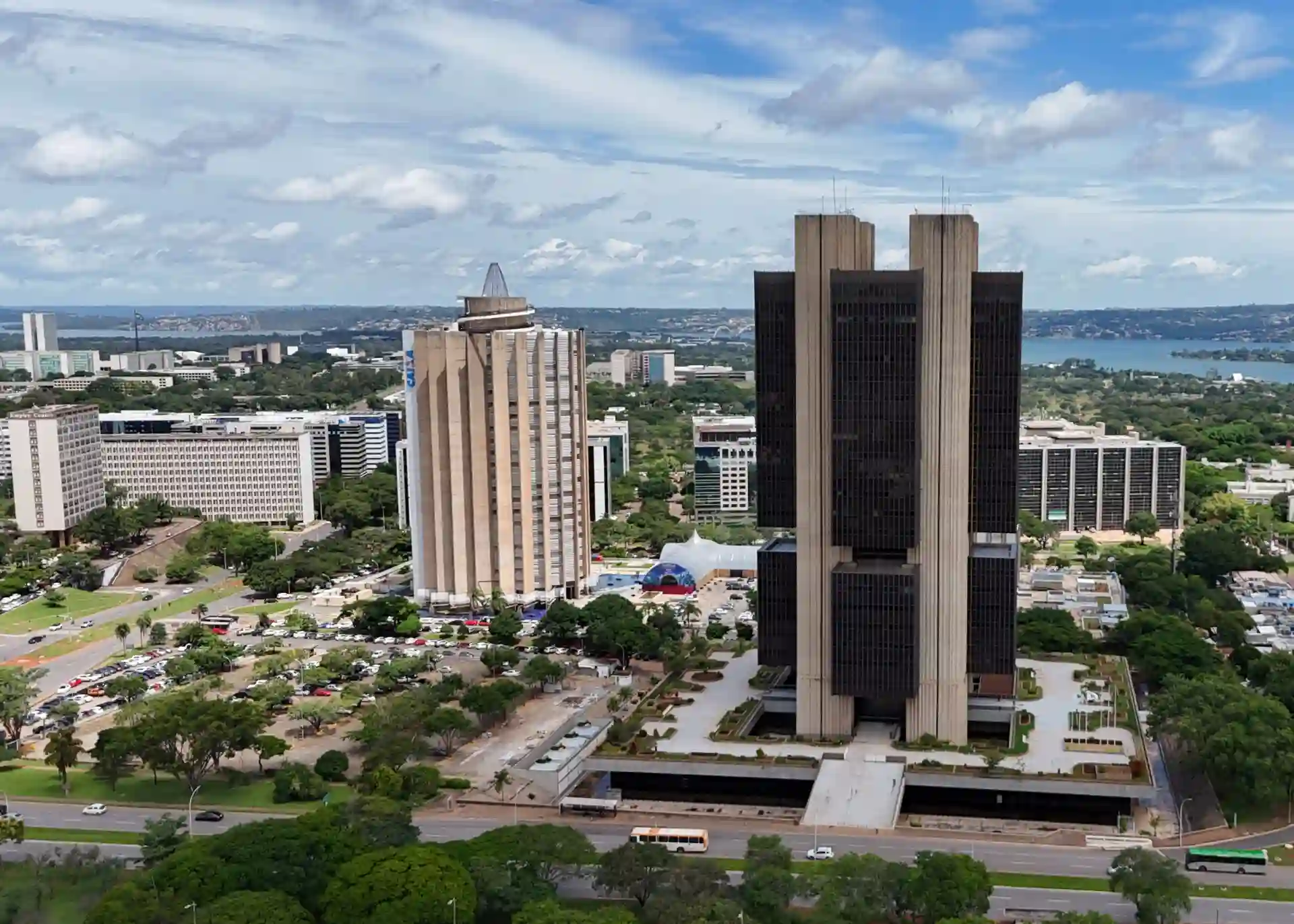 Vista panorâmica de um centro urbano com prédios elevados e áreas verdes ao redor, sob um céu parcialmente nublado, capturada de um drone ou altura elevada.