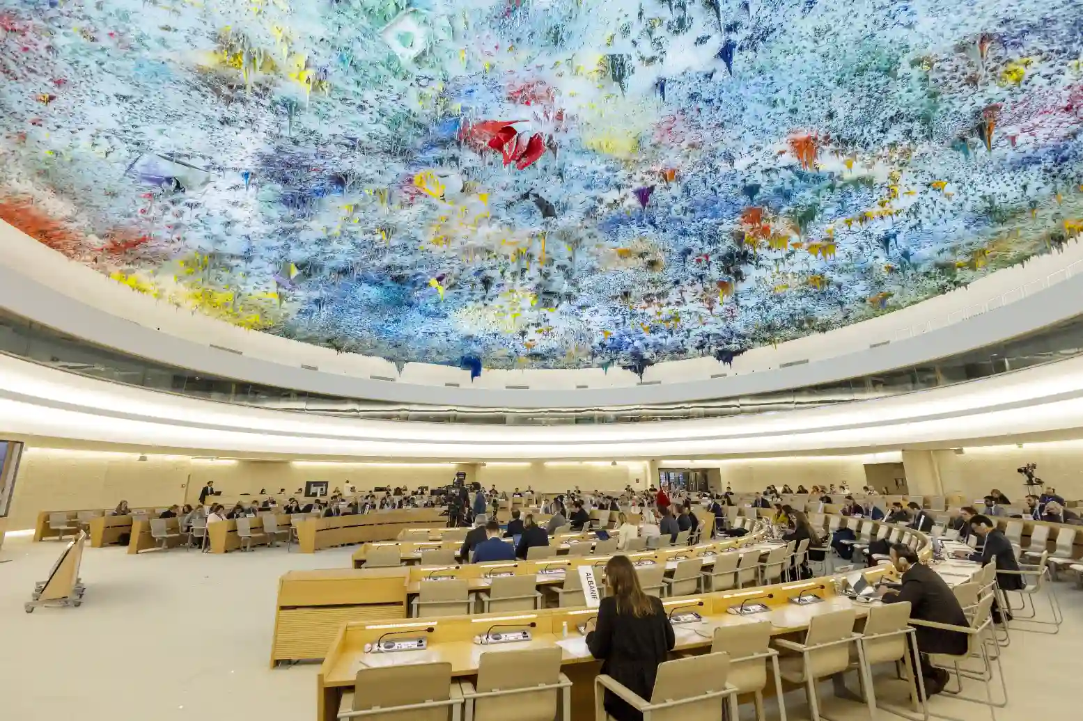Imagem da Conferncia de Diplomata ou evento oficial em uma grande sala com teto artístico colorido e assentos ocupados por pessoas. Ambiente formal e organizado.