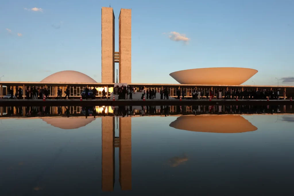 Imagem do Congresso Nacional em Brasília com suas torres emblemáticas e espelho d'água refletindo a arquitetura futurista ao pôr do sol.