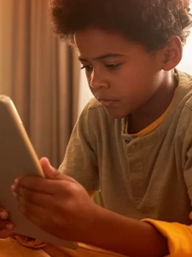 Rapaz jovem usando tablet de forma concentrada em um ambiente doméstico, enquanto uma mulher ao fundo trabalha na cozinha, ambiente acolhedor.