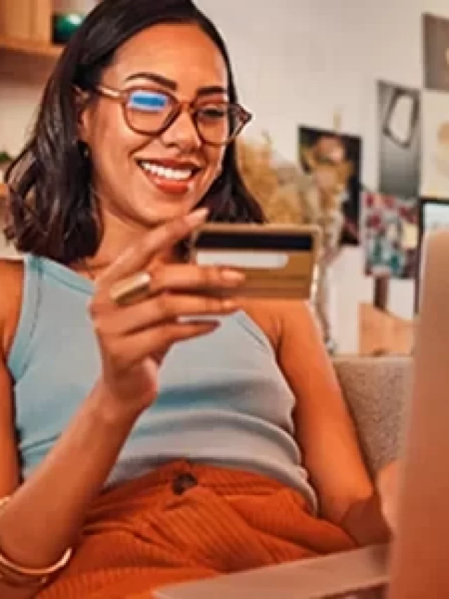 Mulher sorridente usando laptop e cartão de crédito em casa, destacando compras online e facilidade de pagamento na internet, conceito de comércio eletrônico.