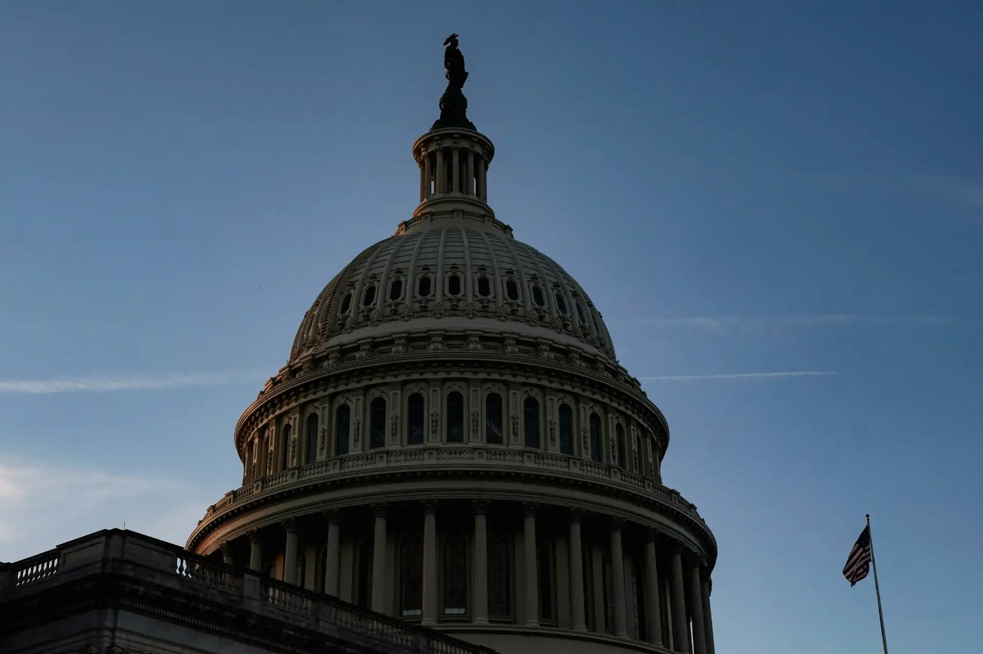 Imagem da cúpula do Capitólio dos Estados Unidos vista ao entardecer, com céu azul e uma bandeira americana ao lado. Arquitetura imponente e histórica.