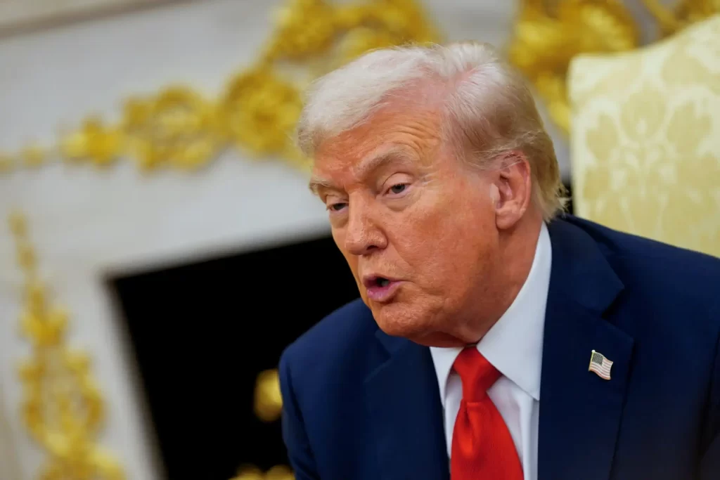 Imagem de Donald Trump, ex-presidente dos Estados Unidos, falando em um ambiente formal, com expressão séria e cabelos brancos. Fundo decorado com detalhes dourados.