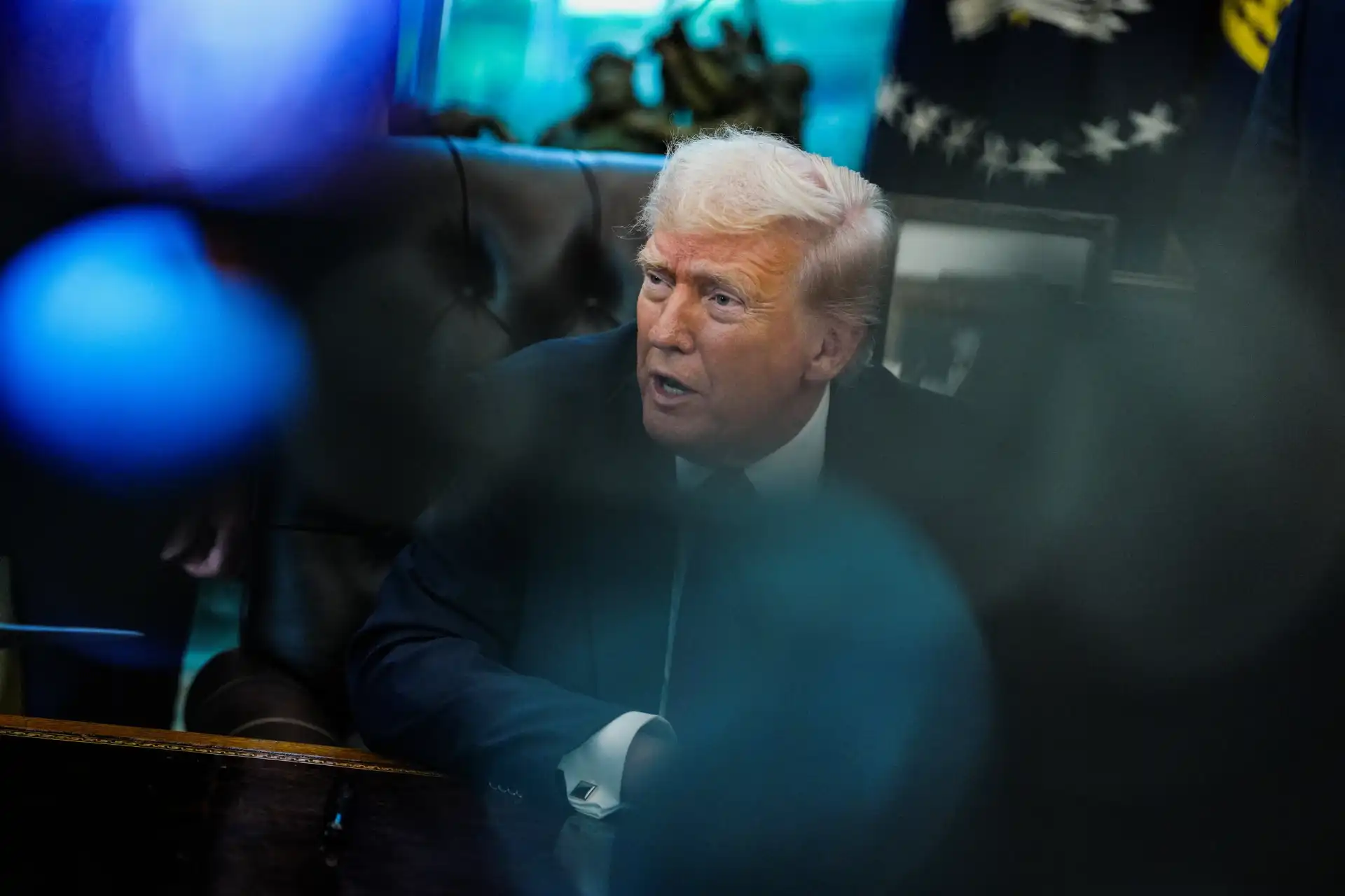 Imagem de Donald Trump em uma reunião, com expressão séria, cercado por pessoas e câmeras, em um ambiente de alta atenção aos detalhes.