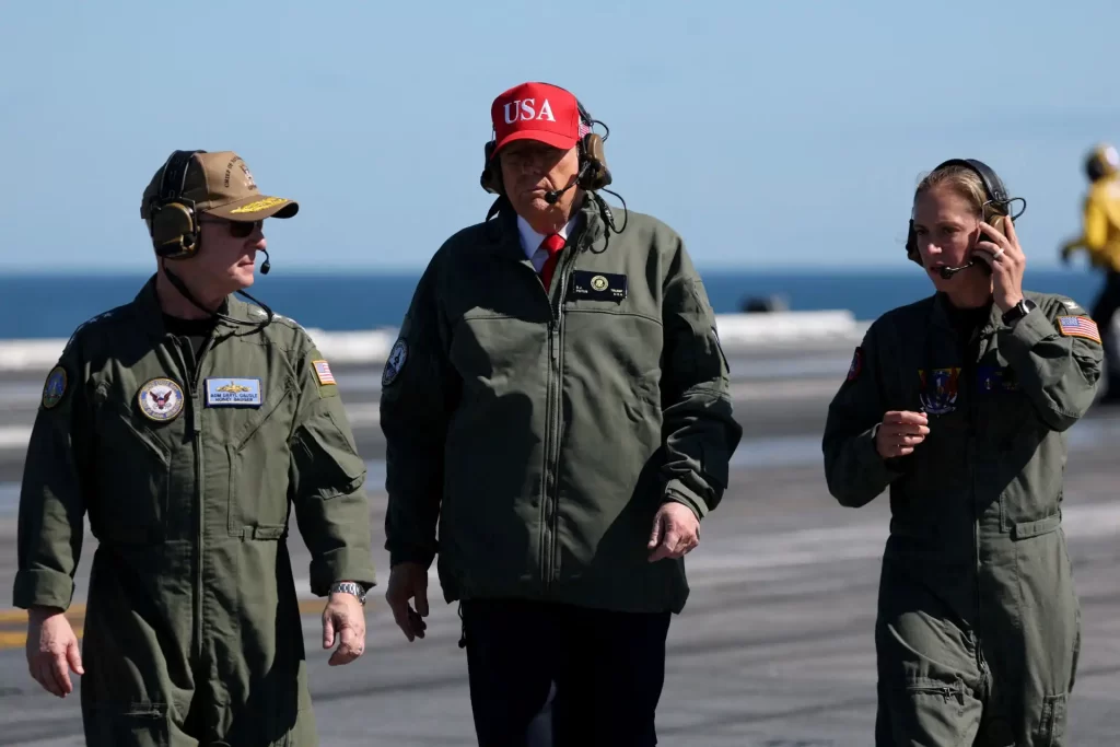 Imagem de Donald Trump em visita a uma base aérea, acompanhado por duas militares. A foto destaca a presença de autoridades americanas e militares durante uma inspeção.