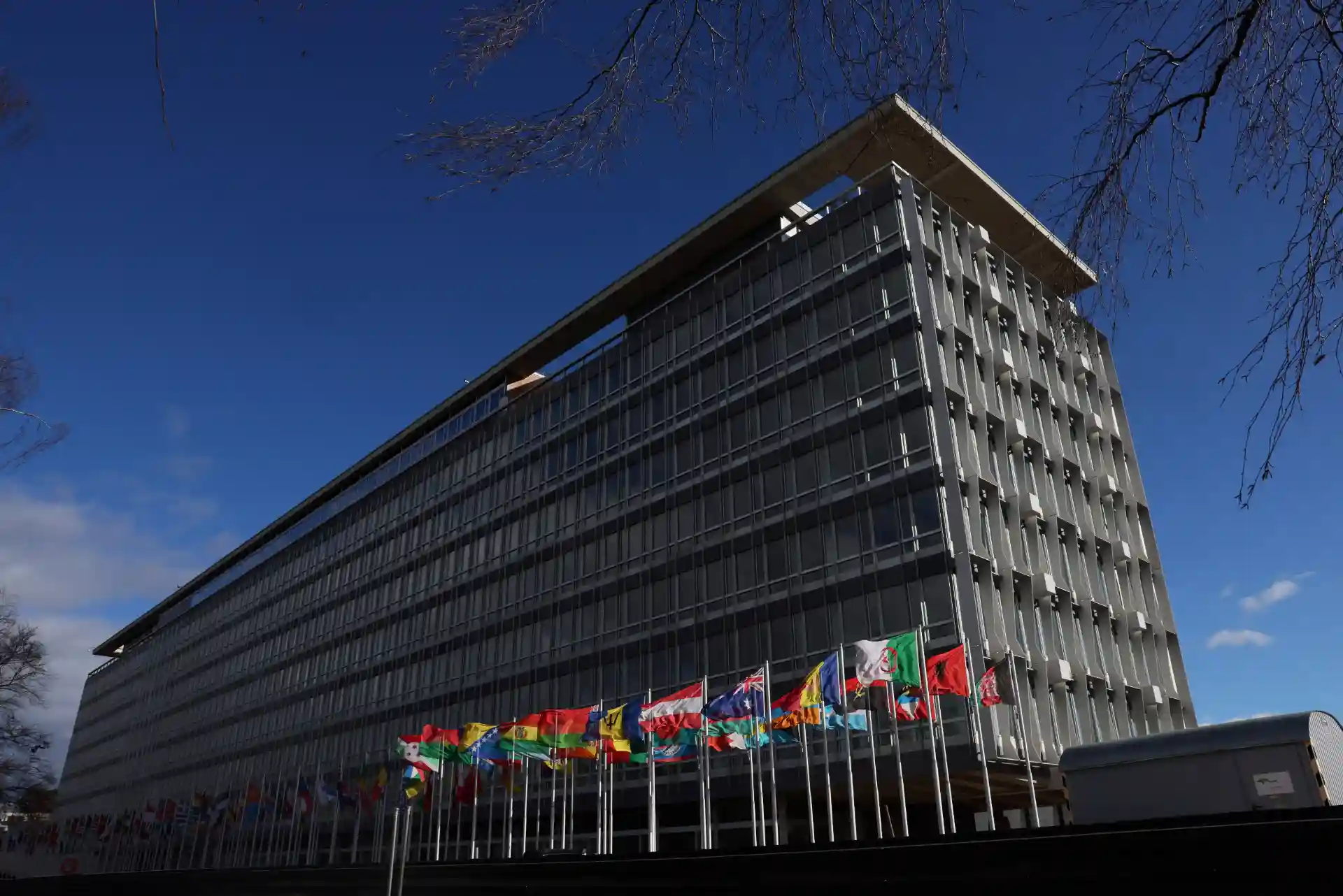 Imagem do edifício do Conselho Europeu, situado em Bruxelas, com várias bandeiras de países membros ao pé, sob céu azul claro.