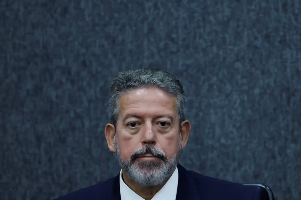 Homem de expressão séria com cabelo grisalho e barba, usando terno escuro, sentado em ambiente formal com fundo azul escuro.