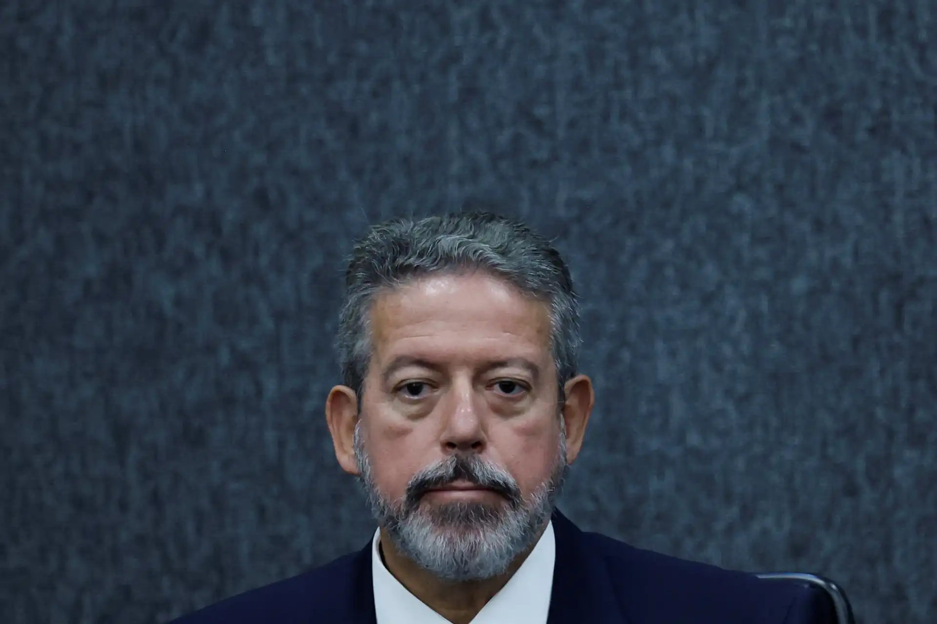 Homem de expressão séria com cabelo grisalho e barba, usando terno escuro, sentado em ambiente formal com fundo azul escuro.