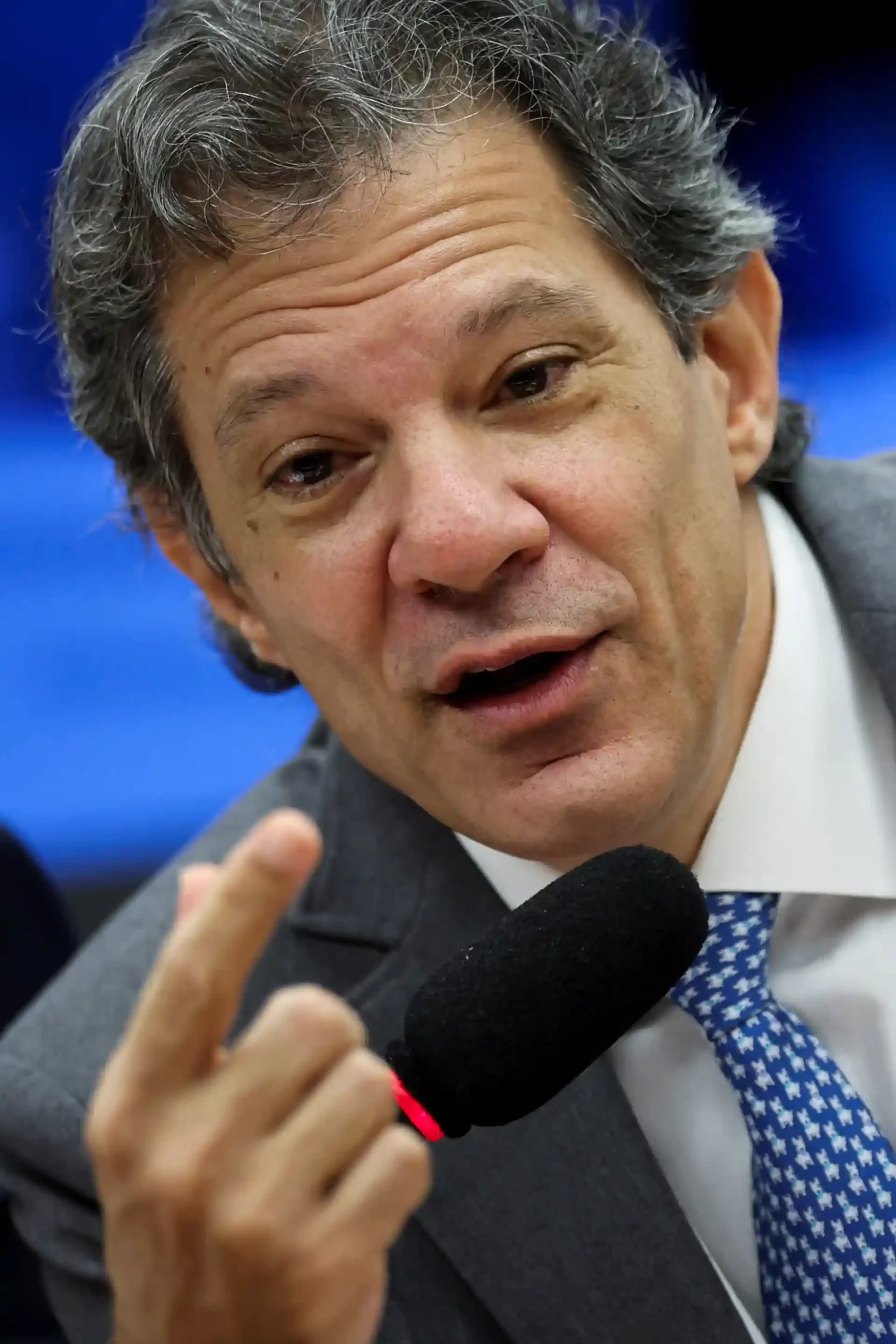 Imagem de um homem falando ao microfone durante uma entrevista ou palestra, usando terno e gravata azul, com cabelo grisalho e expressão séria.