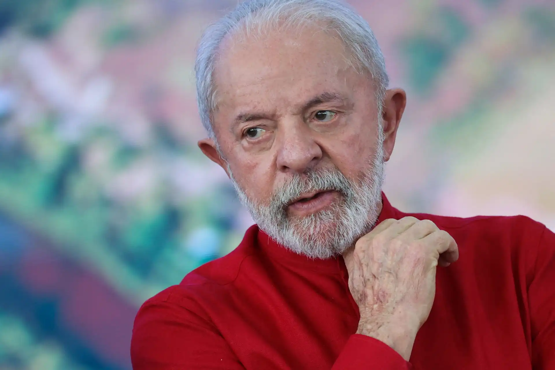 Homem idoso com cabelo grisalho e barba branca, vestindo uma blusa vermelha, expressão pensativa, fundo desfocado com cores suaves.