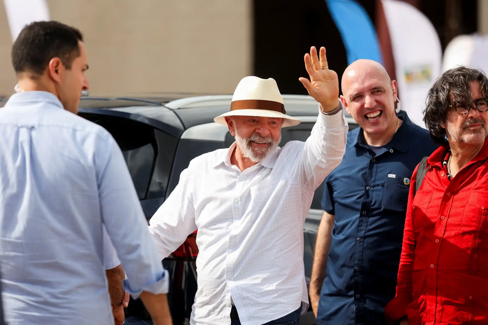 Homem idoso com chapéu de palha e camisa branca, sorrindo e acenando, comemorando um evento ao lado de outros homens em uma reunião ao ar livre.
