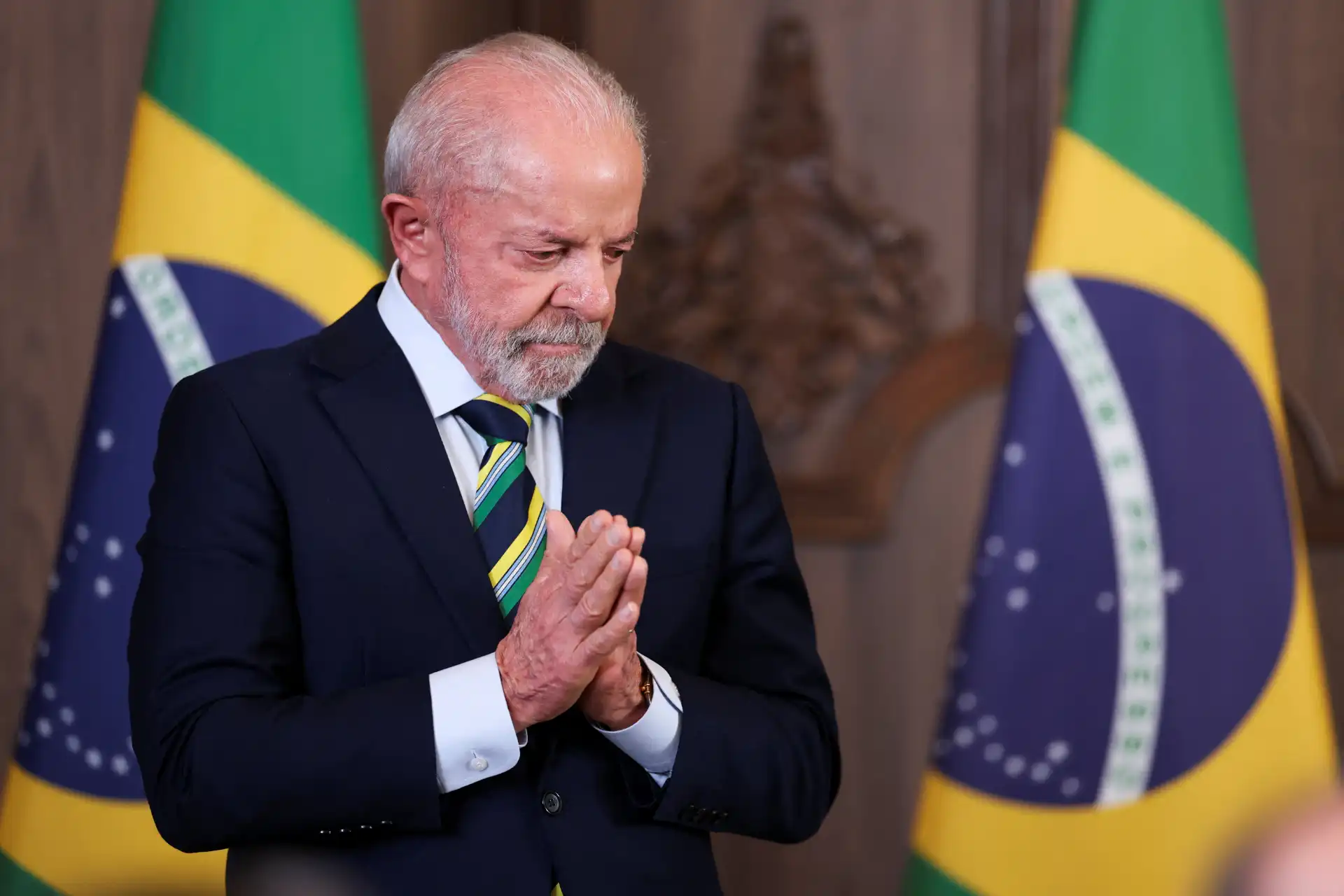 Homem idoso de terno escuro e gravata, com as mãos juntas em oração, ao lado de bandeiras do Brasil, em gesto de fé ou reflexão.