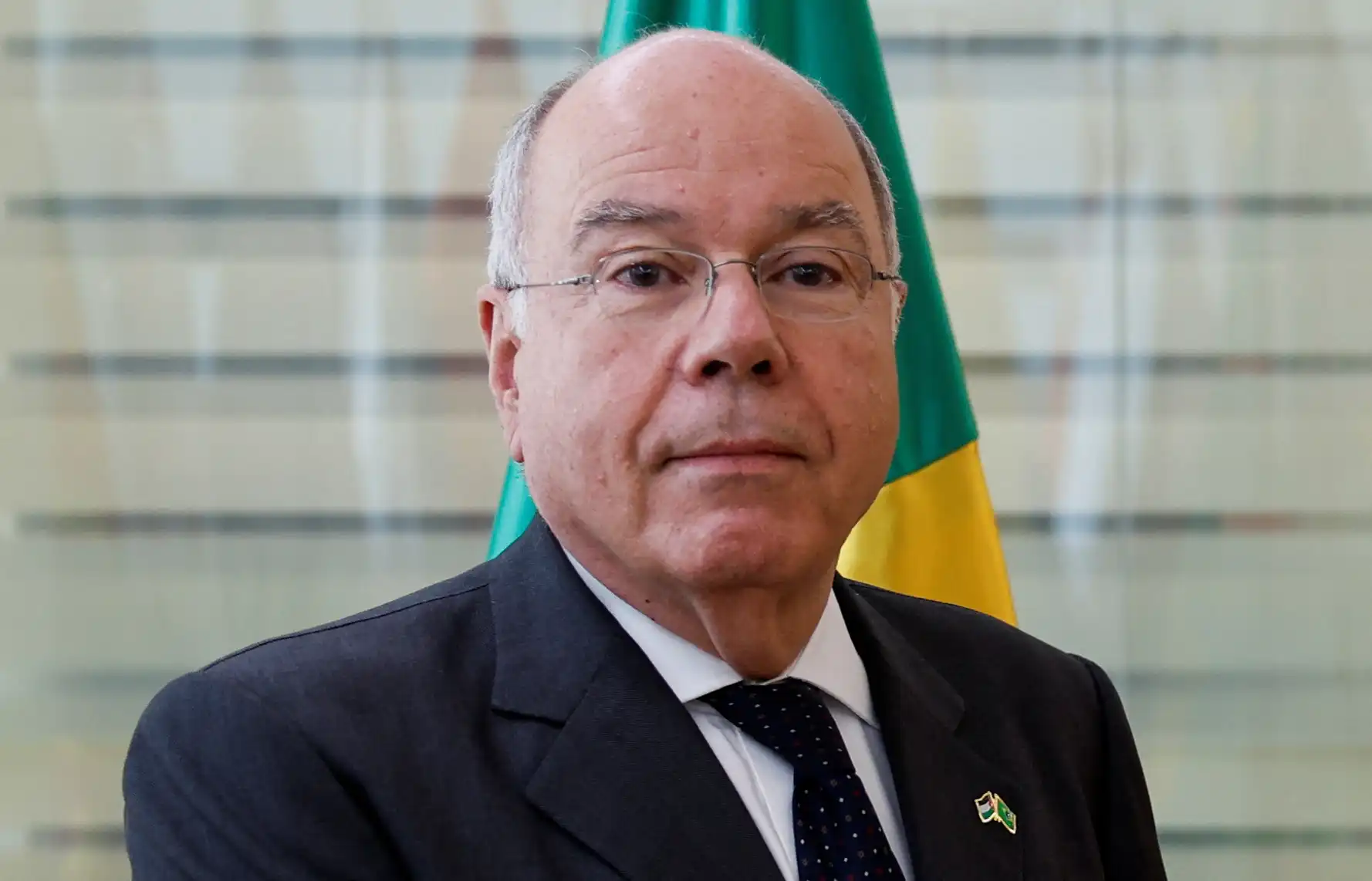 Imagem de um homem de terno, óculos e expressão séria, com bandeira do Brasil ao fundo, representando uma figura política ou líder brasileiro.