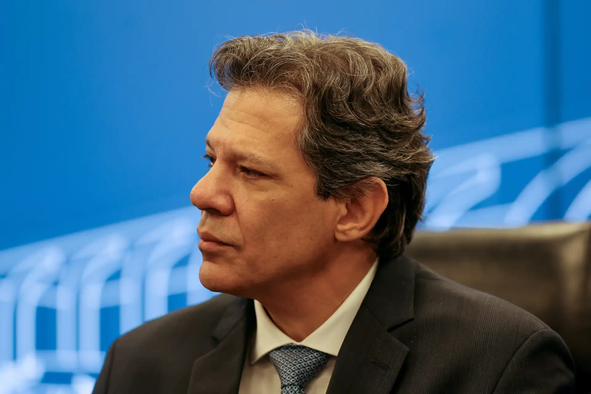 Imagem de um homem de cabelo castanho e grisalho, com expressão séria, usando terno preto e gravata, em ambiente de conferência ou reunião, com fundo azul