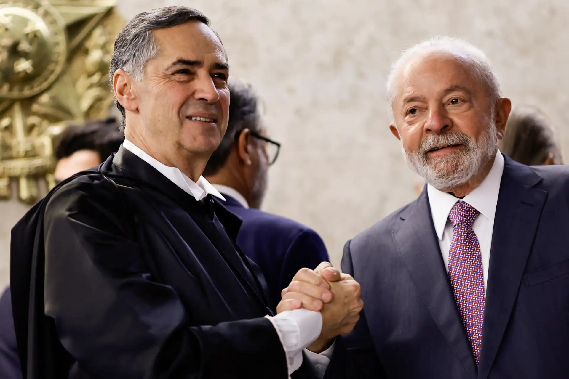 Imagem de dois homens de terno em um evento formal, sorrindo e trocando um aperto de mãos, demonstrando cordialidade em ambiente sofisticado.