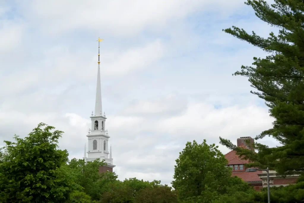 Vista de uma igreja com torre alta e ponteiro dourado em um dia nublado, cercada por árvores verdes e um prédio de cor vermelha ao fundo, em uma paisagem serena.