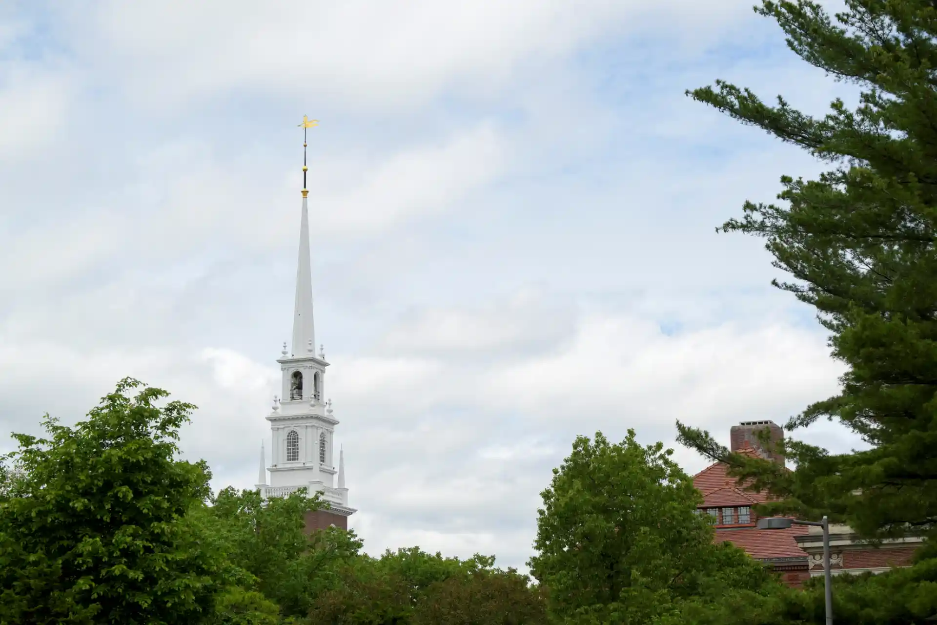 Vista de uma igreja com torre alta e ponteiro dourado em um dia nublado, cercada por árvores verdes e um prédio de cor vermelha ao fundo, em uma paisagem serena.