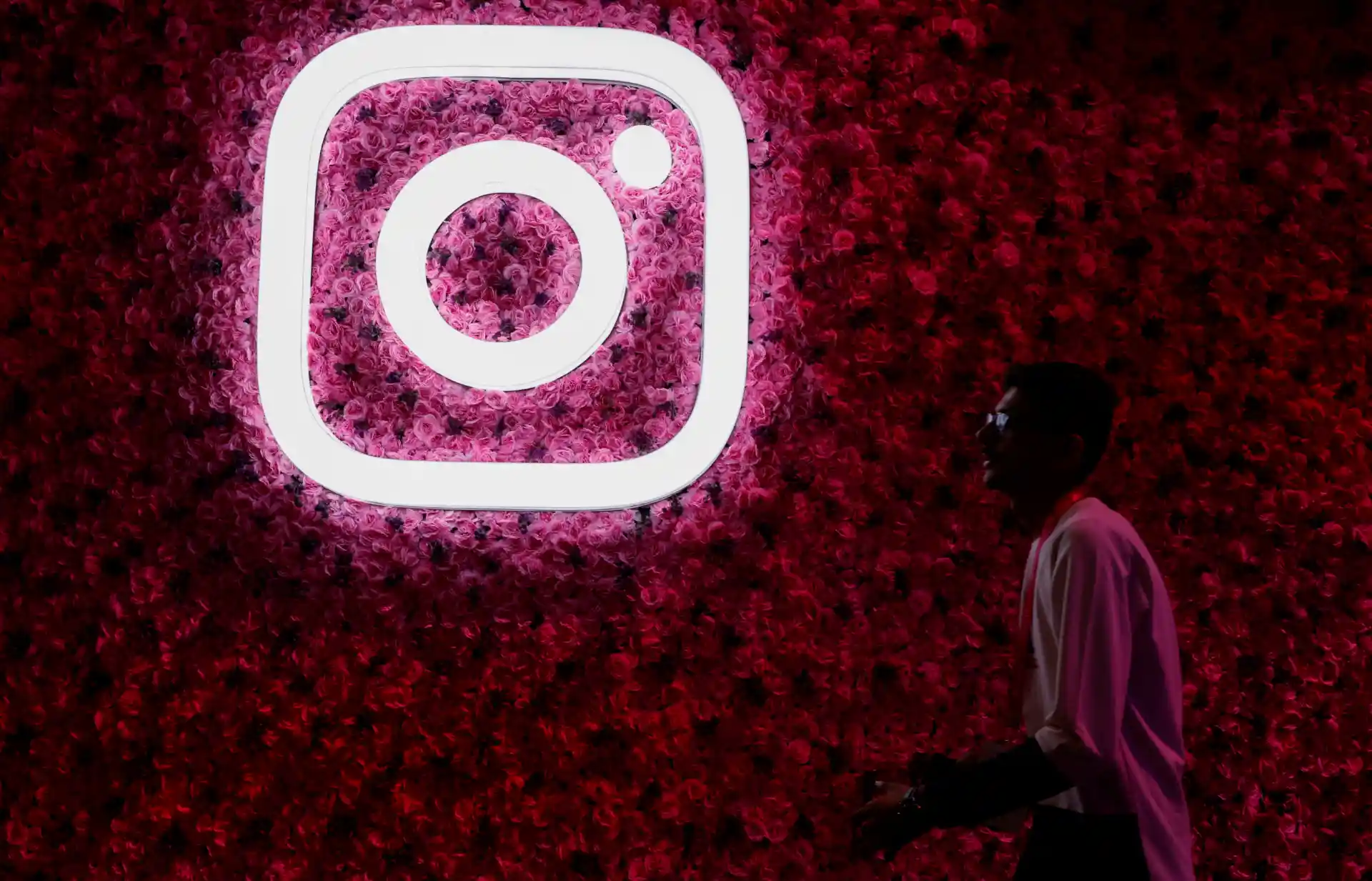 Imagem de um jovem em frente a uma parede decorada com flores vermelhas e o logo do Instagram em neon, destacando-se na foto. Ideal para quem busca conteúdo relacionado à rede social Instagram.
