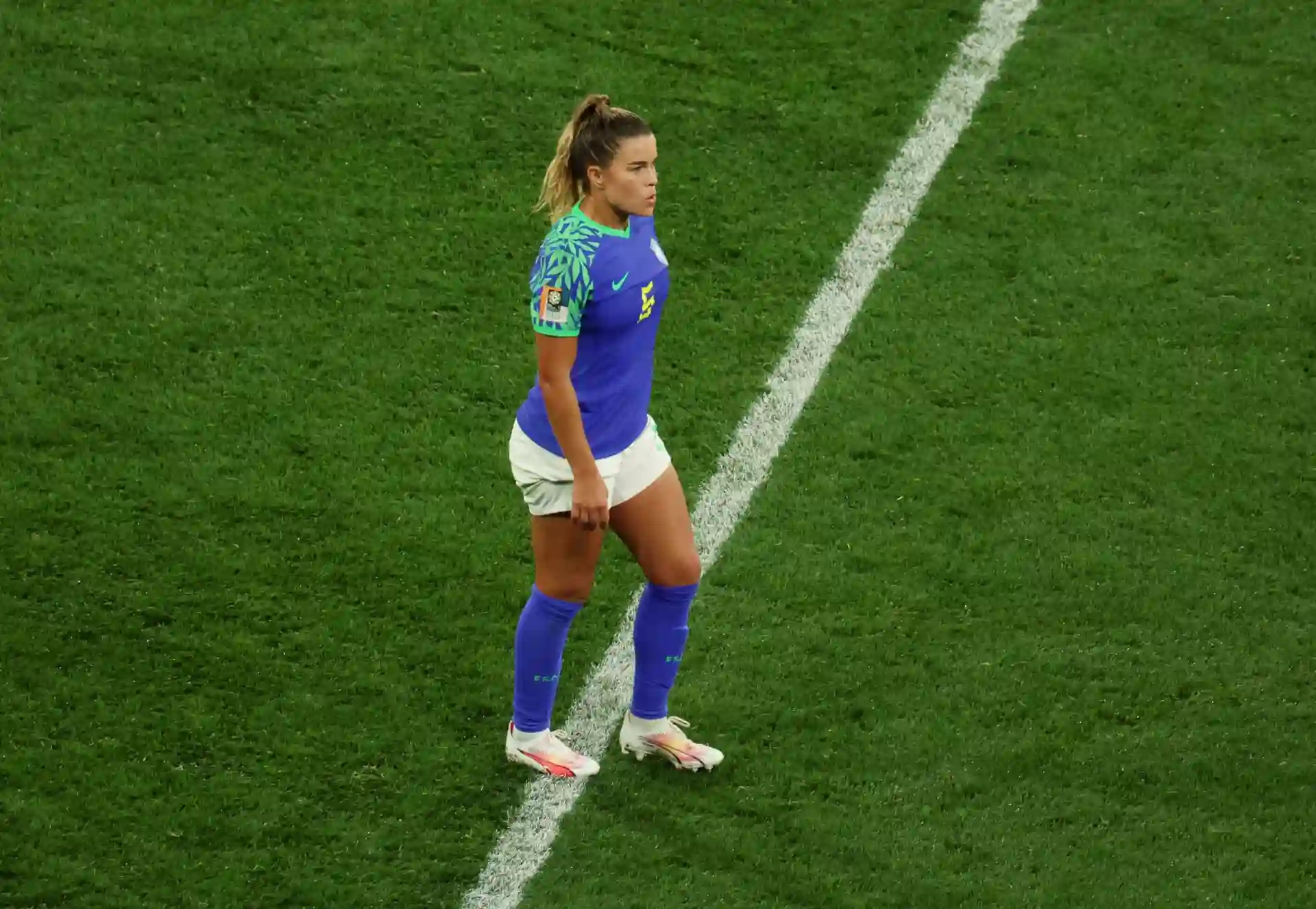 Jogadora de futebol feminina na linha do lance, vestindo uniforme azul e branco, pronta para iniciar a jogada durante a partida.