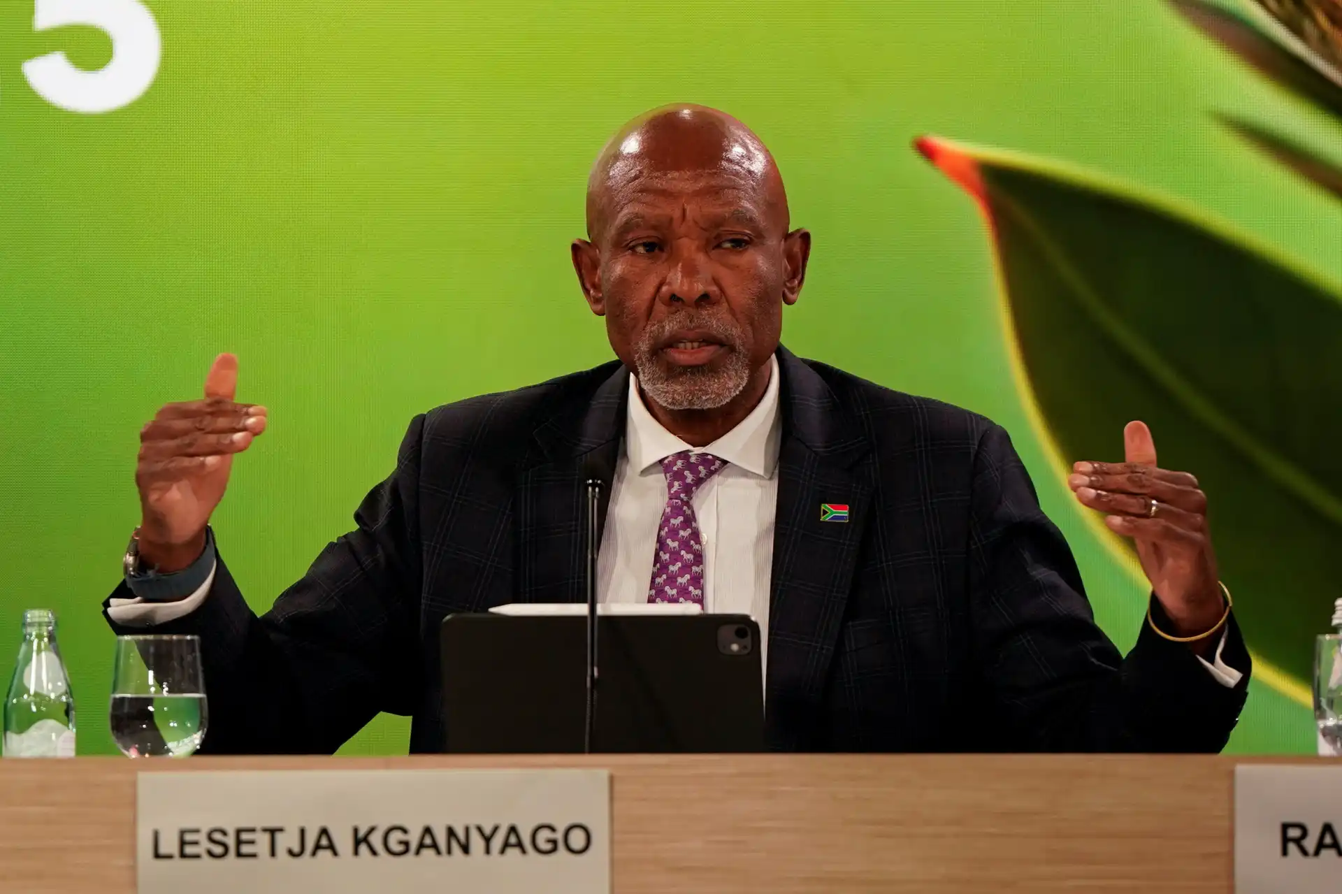 Lesetja KganYago durante uma apresentação ou reunião, sentado atrás de uma mesa com um microfone, vestindo terno e gravata, com fundo verde e alguns objetos na mesa.