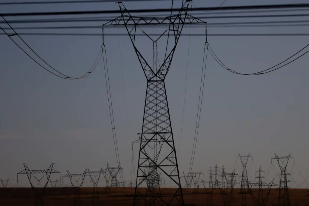 Aneel muda regra para reduzir desconto bilionário a geradores de energia renovável Imagem de linhas de transmissão de energia elétrica com torres em uma paisagem aberta ao pôr do sol.