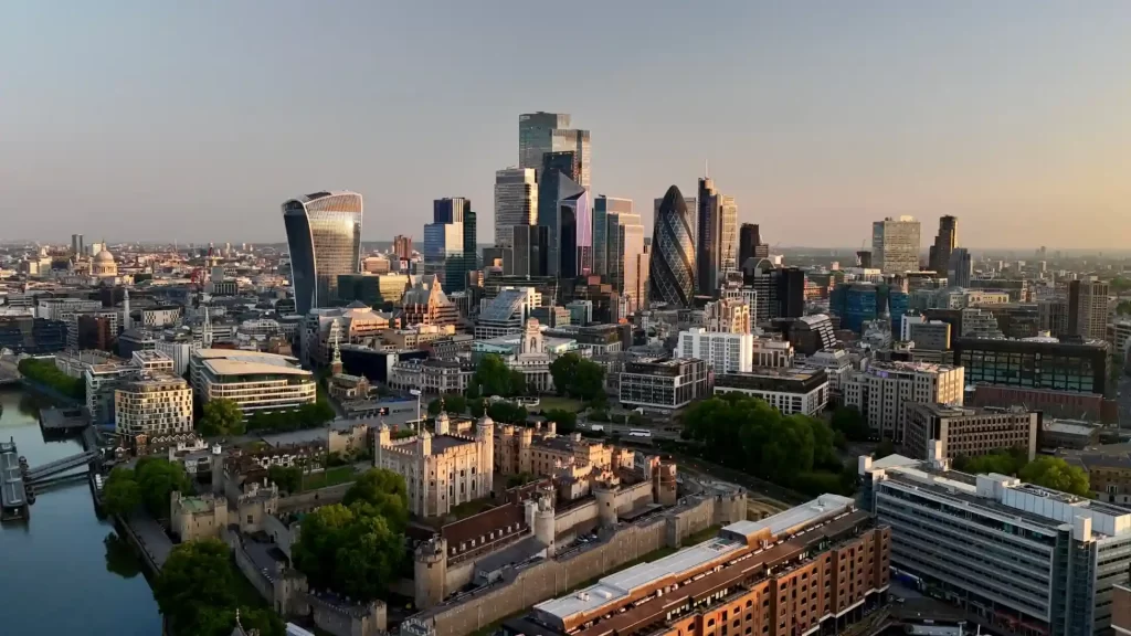 Vista aérea do centro financeiro de Londres com arranha-céus modernos e histórica Torre de Londres ao fundo, durante o pôr do sol.