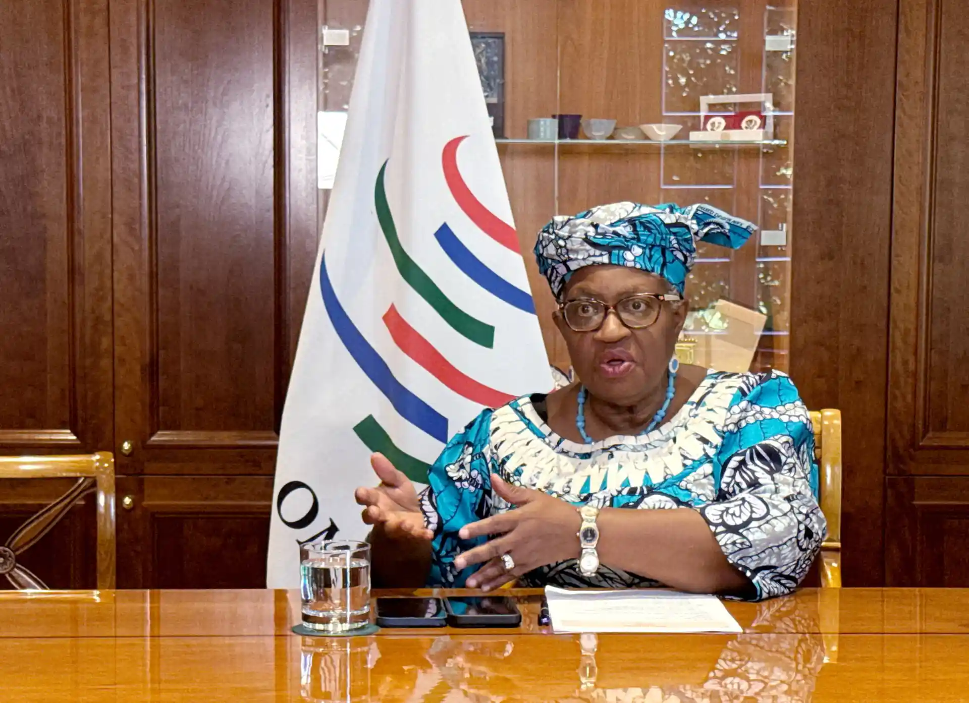 Mulher africana idosa, usando roupas tradicionais e uma headwrap azul, sentada à mesa durante uma reunião oficial, com uma bandeira ao fundo.