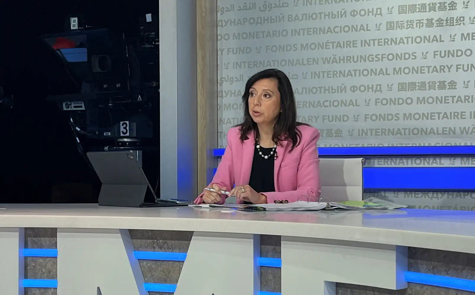 Mulher apresentadora de televisão em estúdio, usando blazer rosa, falando ao vivo sobre economia e finanças internacionais.