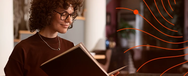 Mulher com cabelos cacheados e óculos lendo um livro em ambiente de casa com elementos decorativos ao fundo, promovendo leitura e bem-estar.