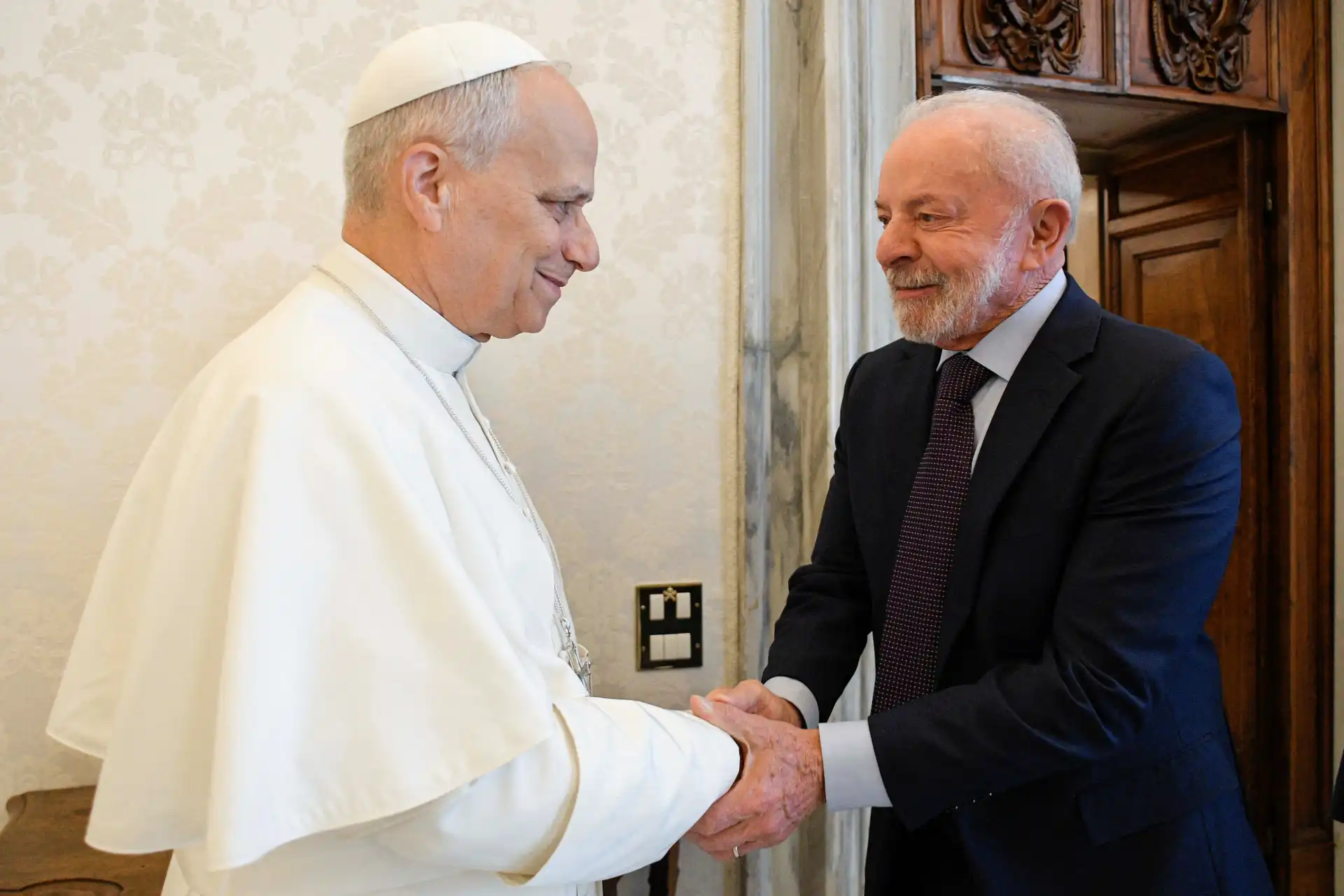 Papa Francisco e um homem idoso sorrindo e se cumprimentando com um aperto de mão em um ambiente elegante, destacando um momento de reunião amistosa