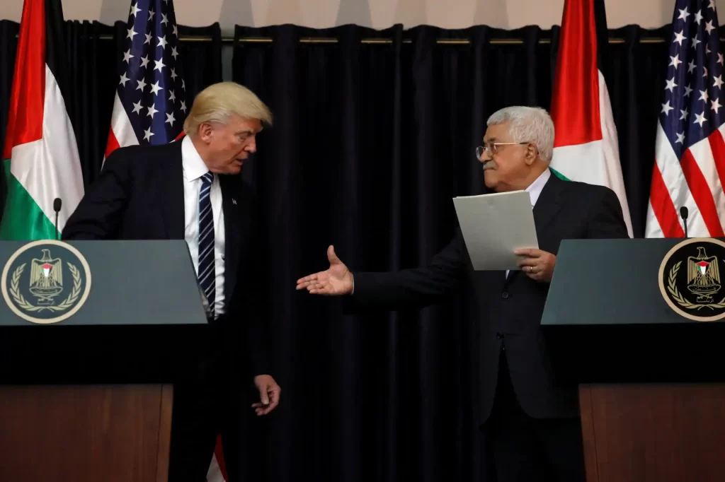 Imagem de duas pessoas, um homem de cabelo loiro e terno escuro, e outro homem com cabelo grisalho, também de terno, trocando uma handshake durante uma entrevista ou conferência, com pódios e bandeiras ao fundo.