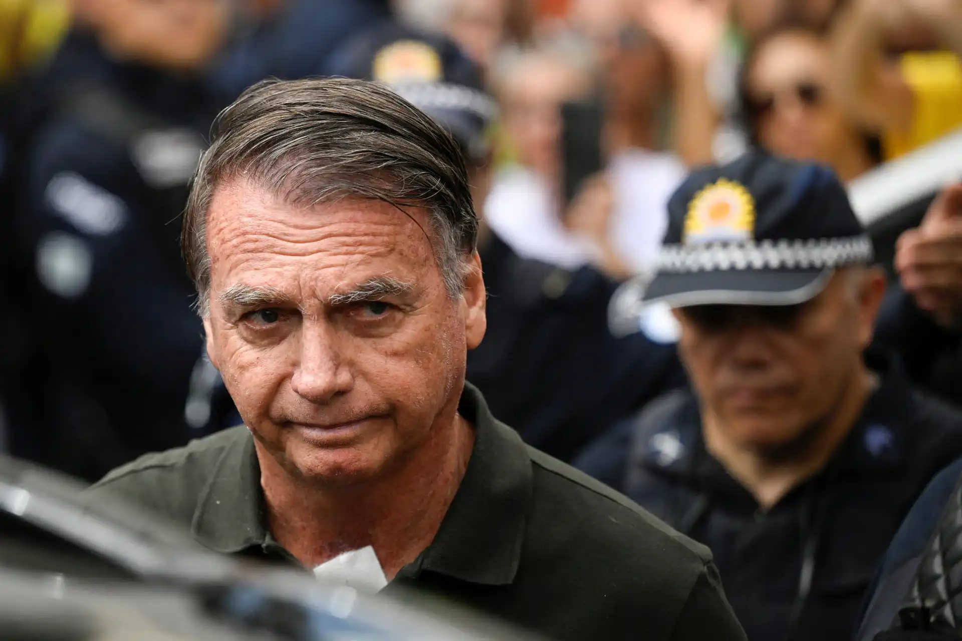 Imagem de uma pessoa masculina de cabelo castanho acinzentado, expressão séria, rodeada por policiais com uniforme e distintivos, em evento público.