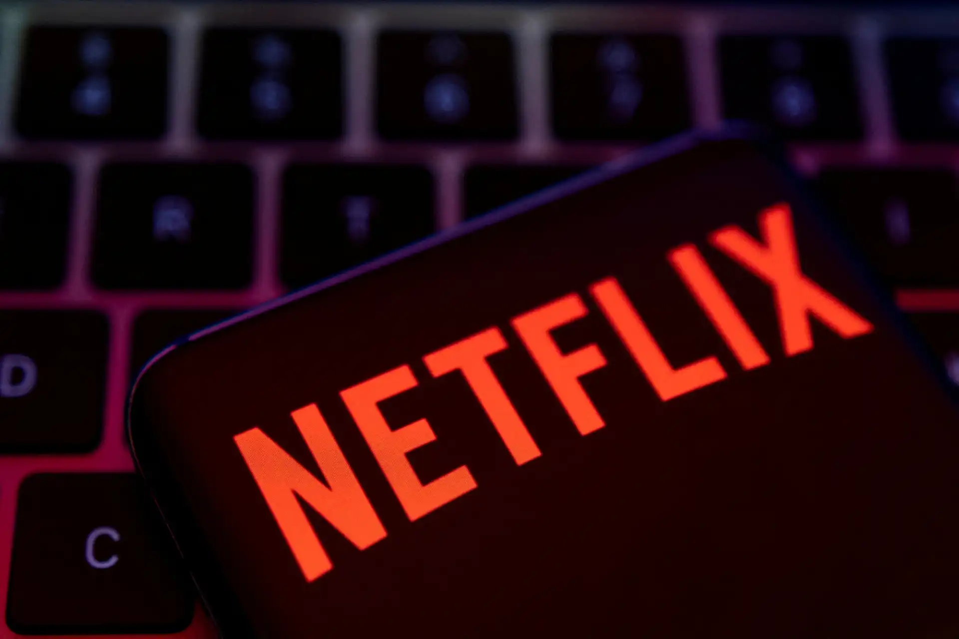 Imagem de um smartphone com o logo do Netflix iluminado em destaque, sobre um teclado iluminado com luz roxa, destacando a palavra Netflix e a experiência de streaming de filmes e séries.