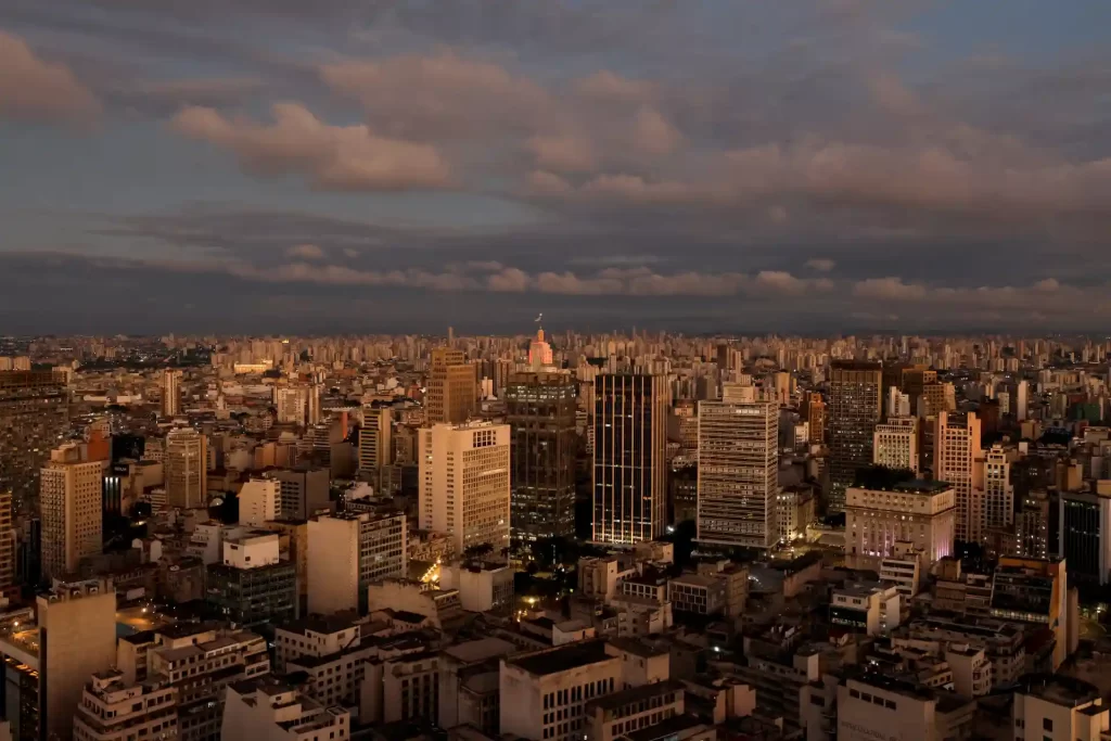 Vista panorâmica da cidade de São Paulo ao entardecer, com predios altos iluminados e céu com nuvens pesadas.