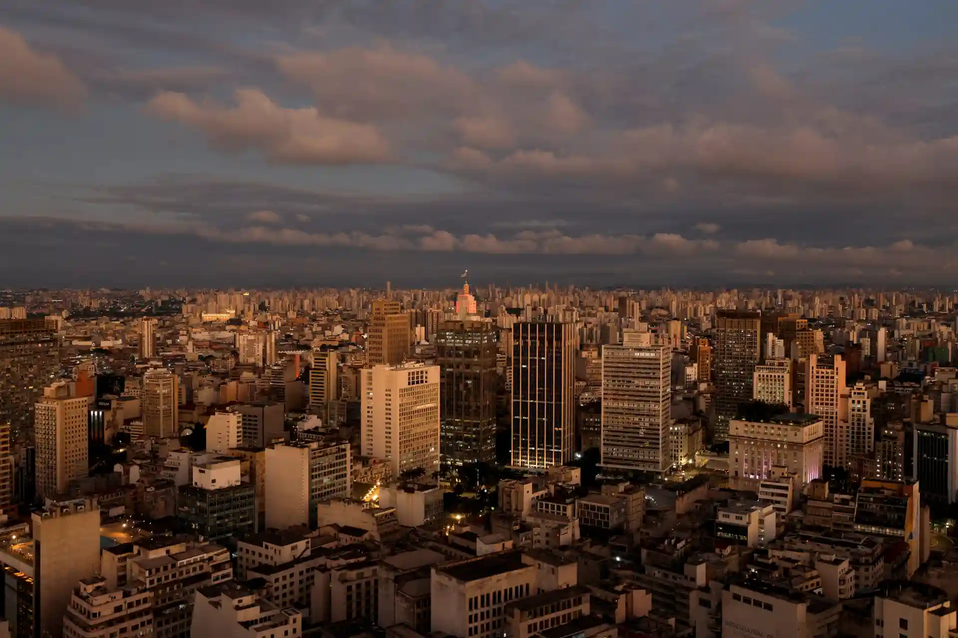 Vista panorâmica da cidade de São Paulo ao entardecer, com predios altos iluminados e céu com nuvens pesadas.