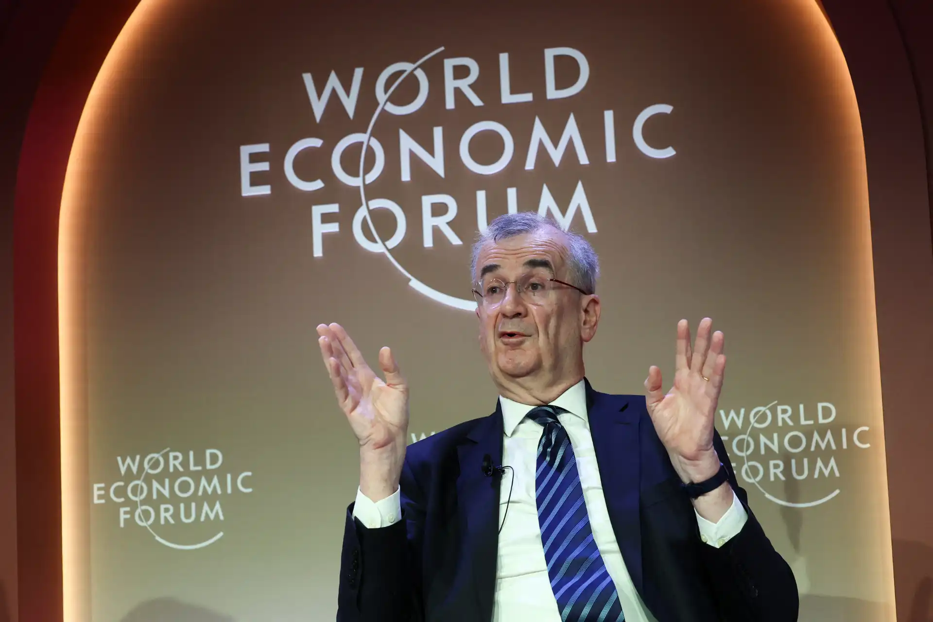 Executivo ou economista falando durante o World Economic Forum, com fundo com o logo do evento. Pessoa vestindo terno, com expressão de explicação e gesticulando com as mãos.