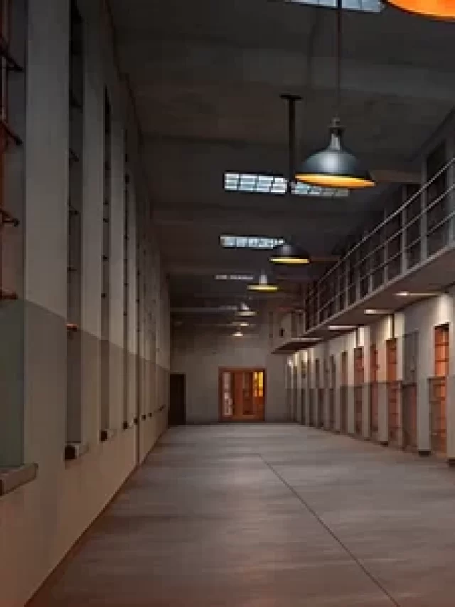 Galeria de celas de prisão com portas de madeira e grades de ferro em ambiente silencioso e bem iluminado com luz natural e iluminação artificial.