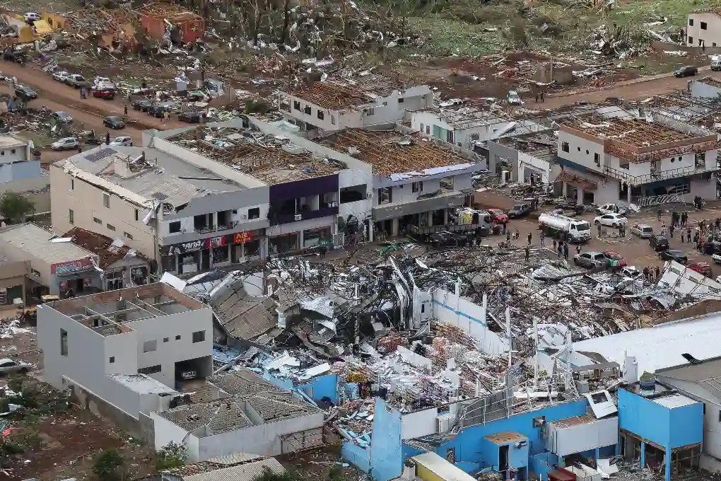 Imagem de uma área urbana devastada por uma tempestade, com prédios destruídos, destroços espalhados e ruas com veículos e pessoas, resultando em uma cena de desastre e destruição.